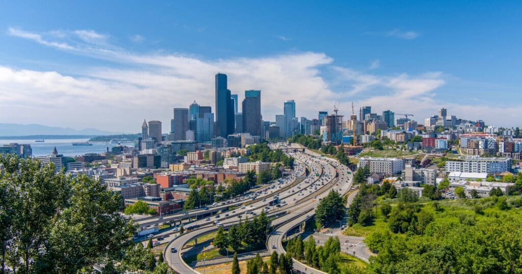 Aerial view of Seattle with skyscrapers, busy highways, and surrounding greenery on a sunny day with clear skies.