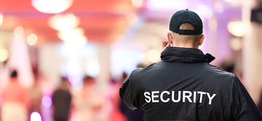 A security officer wearing a black jacket and cap, standing with his back to the camera in a crowded indoor area.