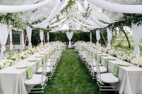 Elegant outdoor private event setup with long banquet tables under a white canopy, decorated for a formal garden event.