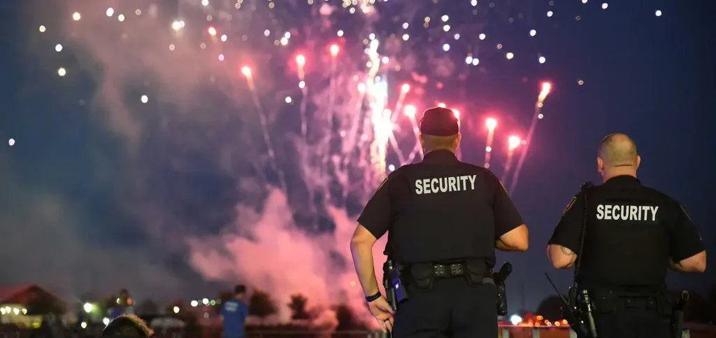 Two security guards standing with their backs to the camera, monitoring a large outdoor event at night while fireworks light up the sky in the background.