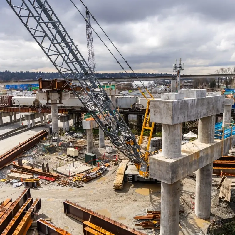 Construction site with large crane lifting steel beams, concrete pillars under construction, and scattered construction materials under cloudy sky.